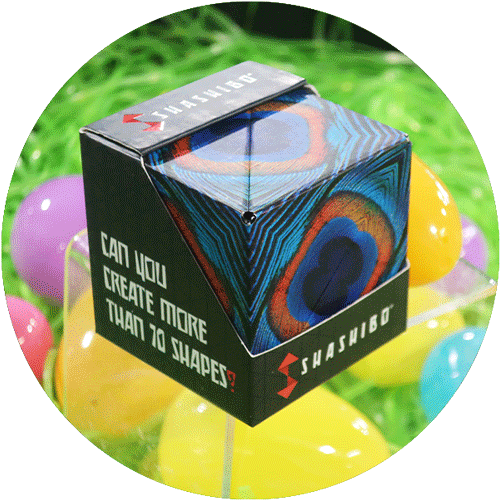 A dark blue, balck and pink cube box puzzle in it's packaging, surrounded by green easter grass and colorful easter eggs.